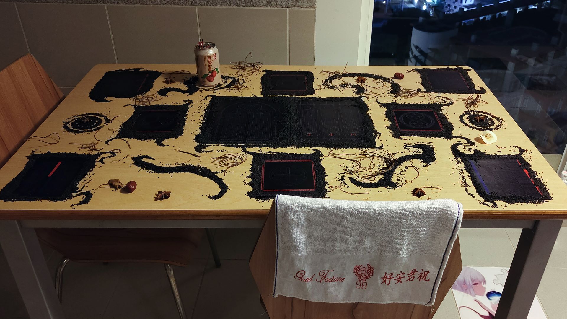 close up of a table with 10 black cards with geometric drawings. they are covered by black sesame seeds at the edges. there is a lychee bottle and small pieces of brown sticks and organic elements