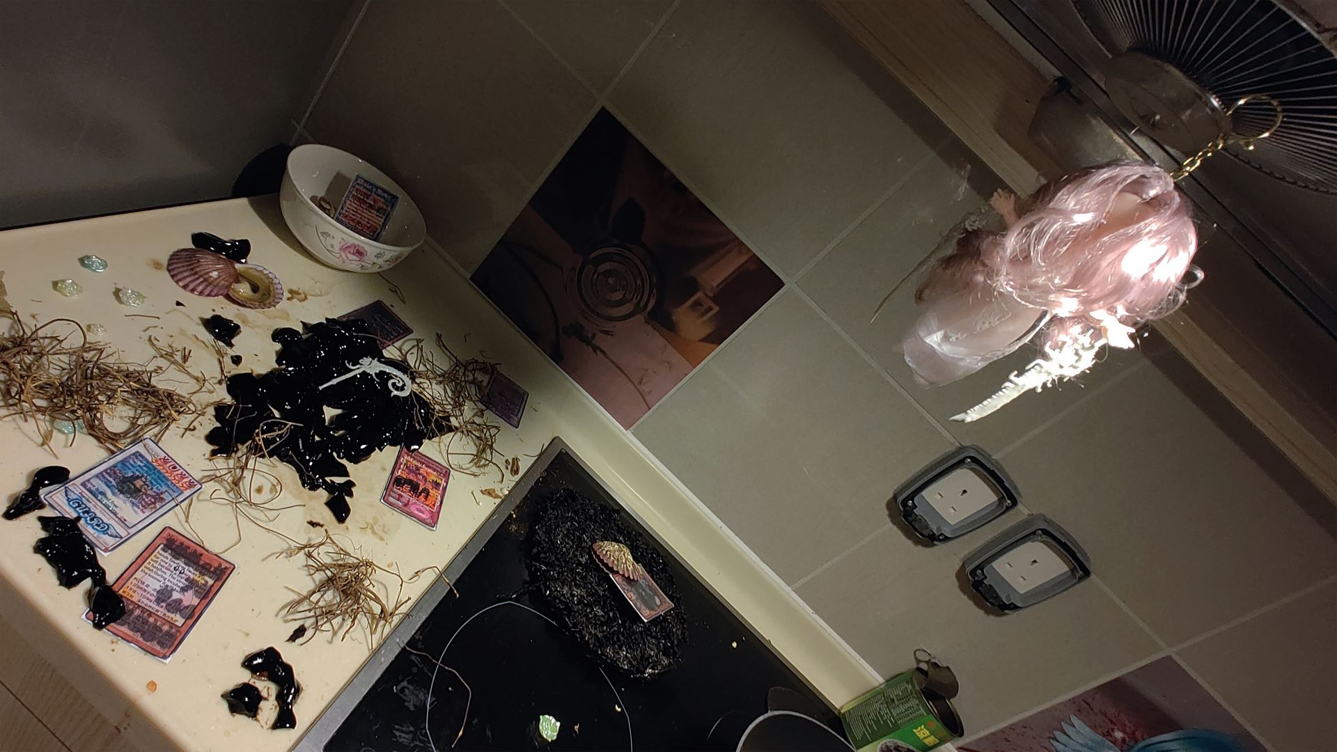 image of the kitchen sink with a pink haired doll with white dress hanging in ventilator. there's black green jelly in counter, yu ji oh cards, shells and small wood twigs