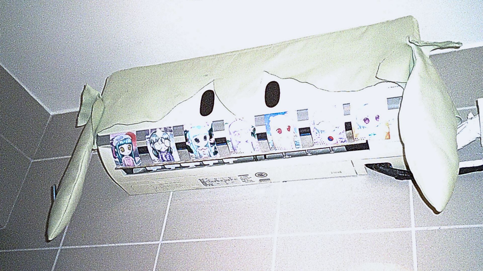 image of an air conditioning machine on the wall with a green cover. the cover is green with ears. it has black eyes making the air conditiong machine look like a doll. there are a series of 7 small images of waifus as teeth.